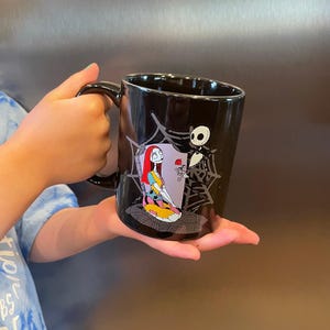 May include: Black ceramic mug featuring Sally and Jack Skellington from The Nightmare Before Christmas. Sally is depicted in her patchwork dress, and Jack is next to her. The mug has a black handle and a glossy finish.
