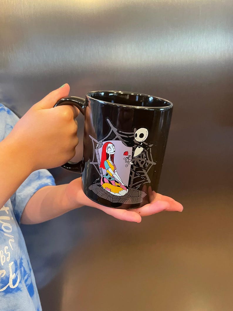 May include: Black ceramic mug featuring Sally and Jack Skellington from The Nightmare Before Christmas. Sally is depicted in her patchwork dress, and Jack is next to her. The mug has a black handle and a glossy finish.