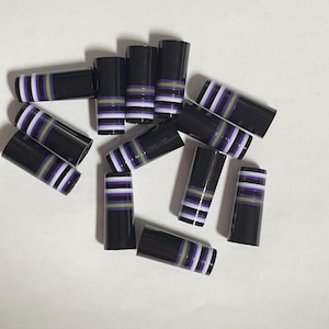 May include: A collection of black cylindrical objects with horizontal stripes of white, purple, and green. The objects are scattered across a white surface, showcasing their decorative design.