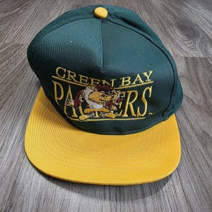 May include: A dark green baseball cap with a yellow brim and a yellow button on top. The front features embroidered yellow text that reads GREEN BAY PACKERS with a graphic of the Tasmanian Devil character positioned in the center.