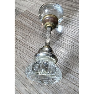 May include: A door knob set featuring a clear, spherical glass knob and a clear, fluted glass knob connected by a square metal spindle. The hardware includes brass-toned collars and a silver-toned base, showcasing a combination of smooth and ribbed textures.