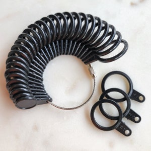 May include: A set of black plastic ring sizers arranged in a circular fan shape on a thin metal cable, with three additional individual black ring sizers detached and lying nearby, each featuring numerical size markings embossed on the small tabs.
