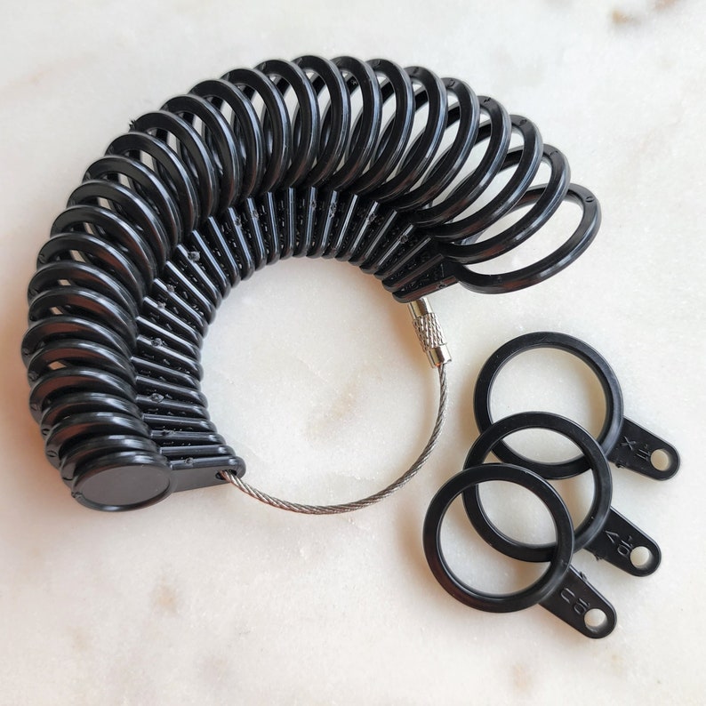 May include: A set of black plastic ring sizers arranged in a circular fan shape on a thin metal cable, with three additional individual black ring sizers detached and lying nearby, each featuring numerical size markings embossed on the small tabs.