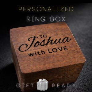 May include: A square wooden ring box with a dark brown finish. The lid is engraved with the text TO Joshua with LOVE in a combination of script and block lettering. The box is presented as a gift-ready item for storing jewelry.