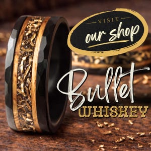 Puede incluir: Un anillo negro martillado con una incrustaci&oacute;n central que presenta vetas de madera y fragmentos met&aacute;licos dorados. El texto "VISIT our shop" aparece en un &oacute;valo con borde dorado, y "BULLET WHISKEY" est&aacute; escrito en letras blancas y doradas.