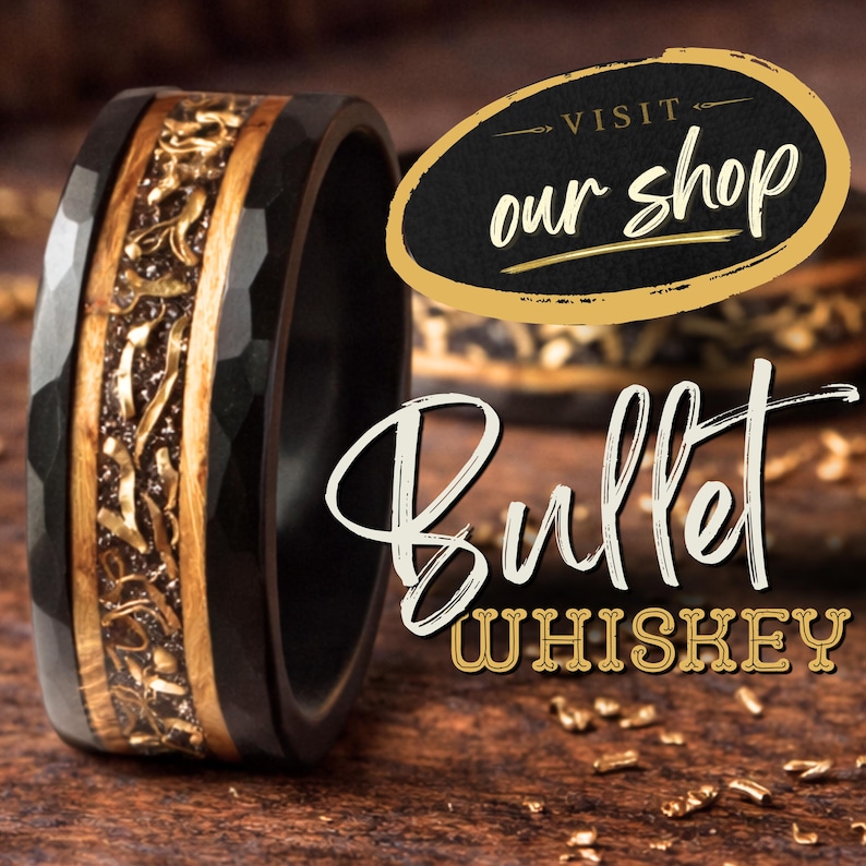 Puede incluir: Un anillo negro martillado con una incrustaci&oacute;n central que presenta vetas de madera y fragmentos met&aacute;licos dorados. El texto "VISIT our shop" aparece en un &oacute;valo con borde dorado, y "BULLET WHISKEY" est&aacute; escrito en letras blancas y doradas.
