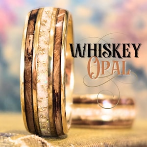 May include: A ring featuring a central band of white opal with gold flecks, flanked by two dark brown wood inlays and polished gold edges. The text "WHISKEY OPAL" is displayed in black and copper lettering to the right of the upright ring.
