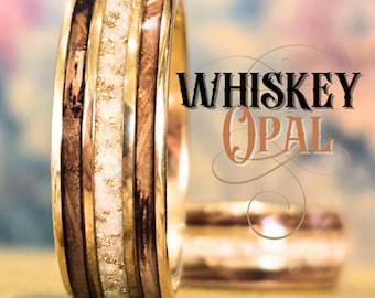 May include: A ring featuring a central band of white opal with gold flakes, flanked by two dark brown wood inlays and polished gold metal edges. The text WHISKEY OPAL is displayed in a stylized black and copper font next to the ring.