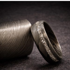 May include: A close-up of a Damascus steel ring with a row of small, clear stones set into the band. The ring is next to a matching cylindrical object, both with a unique wood grain pattern. The background is a dark gray.