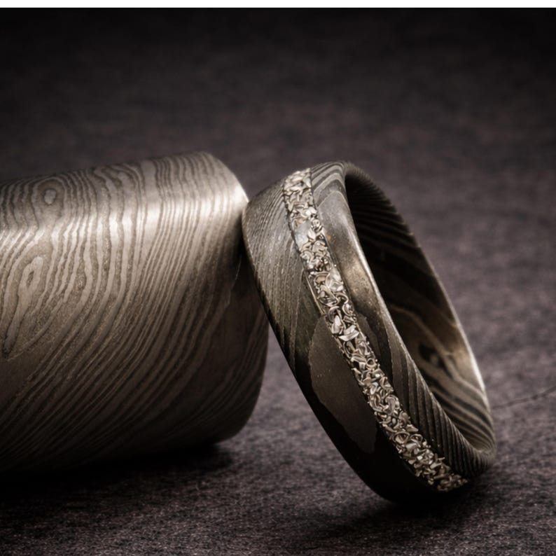May include: A close-up of a Damascus steel ring with a row of small, clear stones set into the band. The ring is next to a matching cylindrical object, both with a unique wood grain pattern. The background is a dark gray.