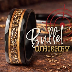 May include: A black hammered metal ring featuring a central inlay of light brown wood and gold-colored metallic shavings. The text "Bullet WHISKEY" is overlaid on the image, accompanied by a red circular target graphic design element.