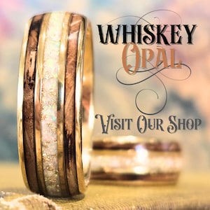 May include: A ring featuring a central band of white opal with gold flecks, flanked by two dark wood inlays and polished gold edges. The text WHISKEY OPAL and VISIT OUR SHOP is displayed in black and orange script next to the ring.