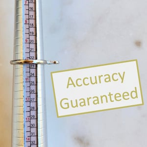 May include: A silver-toned ring positioned on a metal ring mandrel measuring tool. The ring rests at the 22.5 mark on the scale. A rectangular graphic overlay with a white background and gold border displays the text Accuracy Guaranteed.
