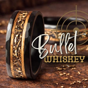 May include: A black hammered metal ring featuring a central inlay of light brown wood and gold-colored metallic shavings. The text "Bullet WHISKEY" is overlaid on the image along with a gold-colored crosshair graphic design element.