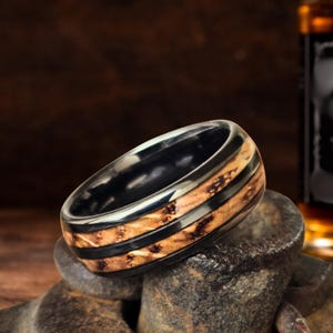 May include: A ring featuring a polished black metal band with a central inlay of textured, light brown wood grain. The design includes two thin black metal stripes running horizontally through the center of the wooden inlay, creating a layered look.