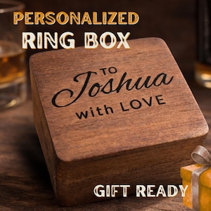 May include: A square, dark brown wooden ring box featuring the engraved text "TO Joshua with LOVE" on the lid. The box has a smooth, polished finish and a hinged design, presenting a clean, simple aesthetic for storing small jewelry items.