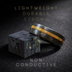 May include: A dark gray ring with a central light brown inlay positioned next to a dark gray cube with a faceted, geometric surface. The text LIGHTWEIGHT, DURABLE, STRONG, and NON CONDUCTIVE is displayed in thin, light-colored capital letters.