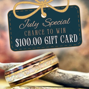 May include: A ring featuring a dark wood grain band with a central inlay of white crushed opal and gold flakes, bordered by thin gold metal edges. Above the ring, a dark teal rectangular tag displays the text July Special CHANCE TO WIN $100.00 GIFT CARD.