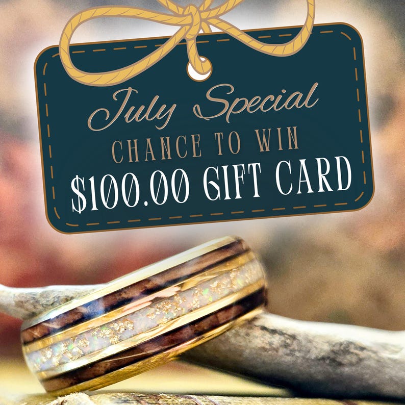 May include: A ring featuring a dark wood grain band with a central inlay of white crushed opal and gold flakes, bordered by thin gold metal edges. Above the ring, a dark teal rectangular tag displays the text July Special CHANCE TO WIN $100.00 GIFT CARD.