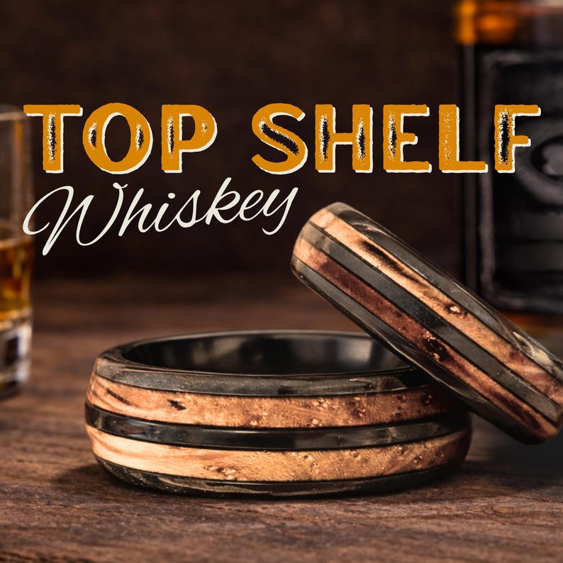 May include: Two rings featuring a layered design with dark black bands and light brown wood grain inlays. The text TOP SHELF Whiskey is displayed in the upper portion of the image, with one ring lying flat and the other resting at an angle against it.