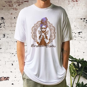 May include: White t-shirt featuring a brown silhouette of Erykah Badu wearing a purple head wrap, with a mandala design behind her. The text "Erykah Badu" is printed below the image.