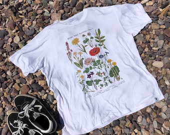 May include: White t-shirt with a print of a botanical illustration of British wild flowers. The print features a variety of flowers in shades of red, yellow, purple, and white. The text "BRITISH WILD FLOWERS - Plate 4" is printed at the top of the illustration.