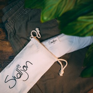 May include: A white drawstring bag with the word "Shellyon" written in black cursive on the front. The bag is lying on top of a pair of green cargo pants.