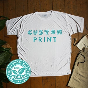 Puede incluir: Camiseta blanca con las palabras "CUSTOM PRINT" en turquesa, con un logotipo circular "FRESH FROM NATURE 100% ORGANIC". La camiseta está sobre una superficie de madera, con pantalones cortos y una bolsa pequeña.
