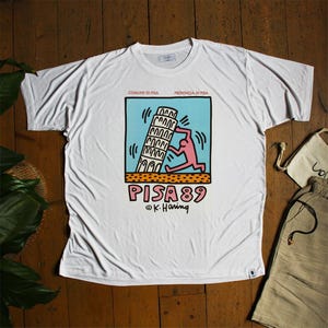 May include: White t-shirt with a graphic print of a person pushing the Leaning Tower of Pisa. The graphic is in black, pink, blue, and yellow. The text "PISA 89" is printed below the graphic. The text "COMUNE DI PISA" and "PROVINCIA DI PISA" is printed above the graphic. The text "© K. Haring" is printed below the text "PISA 89".