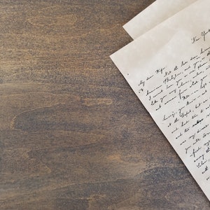 K&ouml;nnte beinhalten: Ein handgeschriebener Brief auf einer braunen Holzoberfl&auml;che. Der Brief ist in der Mitte gefaltet und der obere Teil ist sichtbar. Der Brief ist in Kursivschrift geschrieben und der Text ist teilweise sichtbar. Der Text lautet "Meine liebe Frau...New York...American Babylon...und deine...wie du irgendwo...wo...beeil dich, du beh&auml;ltst et...am Buffet, wette ar...und hatte Zeit zu essen...und zwei ander...mein Abendessen...du wirst aden...zufrieden...emry die...Salen, eitt..."