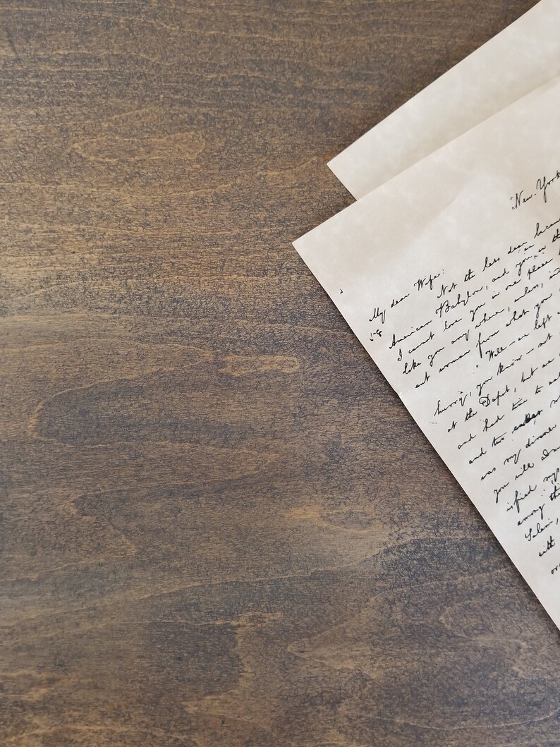 K&ouml;nnte beinhalten: Ein handgeschriebener Brief auf einer braunen Holzoberfl&auml;che. Der Brief ist in der Mitte gefaltet und der obere Teil ist sichtbar. Der Brief ist in Kursivschrift geschrieben und der Text ist teilweise sichtbar. Der Text lautet "Meine liebe Frau...New York...American Babylon...und deine...wie du irgendwo...wo...beeil dich, du beh&auml;ltst et...am Buffet, wette ar...und hatte Zeit zu essen...und zwei ander...mein Abendessen...du wirst aden...zufrieden...emry die...Salen, eitt..."
