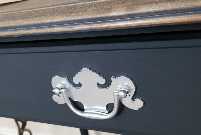 K&ouml;nnte beinhalten: Nahaufnahme einer Schublade mit einem silbernen Schubladenknauf. Der Schubladenknauf hat ein dekoratives Design und ist an einer dunkelgrauen Schublade befestigt.