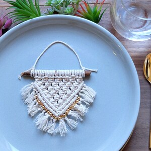 May include: A small white macrame wall hanging with gold beads and a wooden dowel. The hanging is on a light blue plate with a gold fork and knife on a yellow placemat.