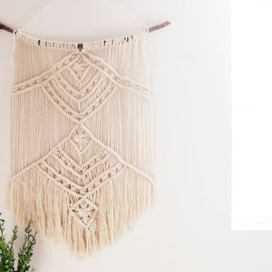 May include: A white macrame wall hanging with a geometric pattern and fringe. The hanging is attached to a branch.