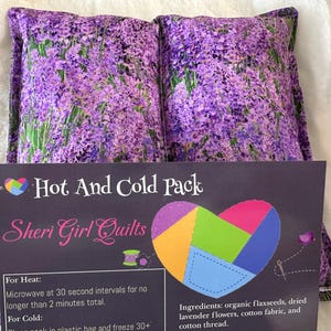 May include: Two lavender-printed hot and cold packs. The packs are rectangular and feature a vibrant purple floral design. A card in front reads "Hot And Cold Pack" and "Sheri Girl Quilts", with instructions and ingredients listed.