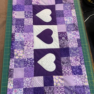 May include: A purple and white patchwork quilt with three white hearts with purple stitching in the center. The quilt is made of various purple fabrics with floral and geometric patterns.