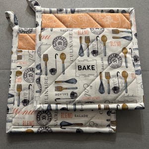 May include: A set of two quilted pot holders with a white background featuring kitchen utensils and the word "BAKE". The pot holders have a peach-colored accent and a loop for hanging.