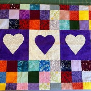 May include: A purple and white patchwork quilt with three hearts in the center. The hearts are made of different fabrics and are surrounded by a border of squares. The quilt is made of a variety of fabrics, including floral prints, solids, and geometric patterns.