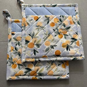 May include: Two square pot holders with a light blue quilted top and a floral pattern of oranges and green leaves on a light blue background. Each pot holder has a fabric loop for hanging.