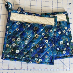 May include: Two square quilted pot holders featuring a dark blue fabric with a white and yellow floral pattern. Each pot holder has a cream-colored top pocket with a subtle geometric print and diagonal stitching. A fabric loop is attached to the corner.