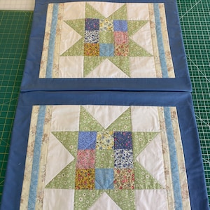 May include: Two blue pillow covers with white fabric and a patchwork star design in shades of green, blue, and pink. The star is made up of smaller squares of fabric with floral patterns.