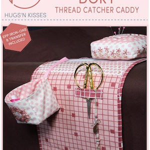 May include: A pink and white thread catcher caddy with a grid pattern, a small pink and white polka dot basket, a pink and white pincushion, and gold scissors. The caddy has a pocket for scissors and a loop for hanging a needle threader. The text "HUNKY DORY THREAD CATCHER CADDY" is at the top of the image.