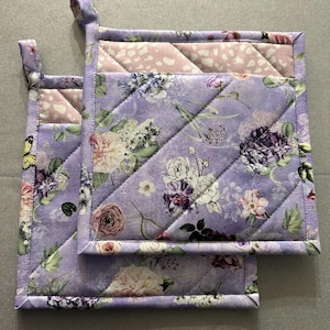May include: Two square quilted pot holders with a floral pattern in shades of purple, pink, and white. The pot holders have a loop for hanging and a diamond-stitched design. The floral pattern includes roses, hydrangeas, and other flowers.