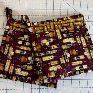 May include: Two burgundy pot holders with a wine-themed pattern. The fabric features illustrations of wine bottles, corks, and glasses in various colors. Each pot holder has a loop for hanging.