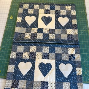 May include: Two blue and white patchwork fabric placemats with white heart cutouts. The placemats are made of a variety of blue and white fabrics, including paisley and polka dots.