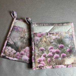 May include: Two square pot holders with a floral print featuring pink flowers and green foliage. The pot holders have a quilted design and a loop for hanging. The background is a muted gray.