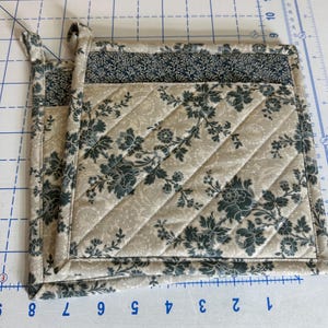 May include: A square quilted pot holder featuring a beige background with a dark green floral pattern. The fabric has diagonal stitching lines and a solid dark green floral border at the top. A small fabric loop is attached to the top corner.