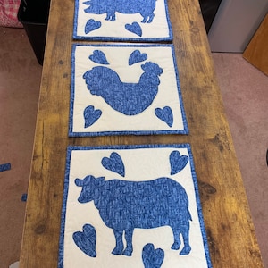 May include: Three white fabric placemats with blue embroidered designs of a pig, rooster, and cow. Each design is surrounded by blue hearts.