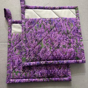 May include: Two quilted pot holders with a vibrant purple floral pattern, featuring a lavender field design. The pot holders have a white dotted border and a loop for hanging. The quilting creates a diamond pattern across the surface.