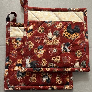 May include: Two quilted pot holders with a rooster and sunflower pattern. The main fabric is a deep red with a repeating design of roosters, sunflowers, and other floral elements. The top section is a cream-colored fabric with a quilted pattern.